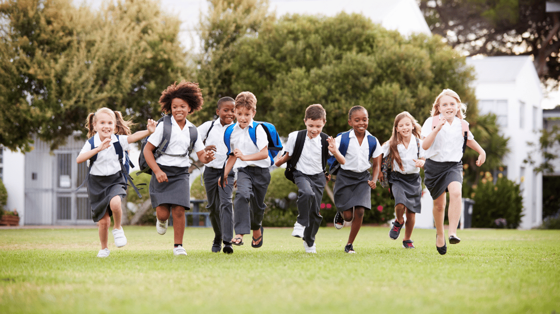 Photo of children in school uniforms running across a green lawn with backpacks on